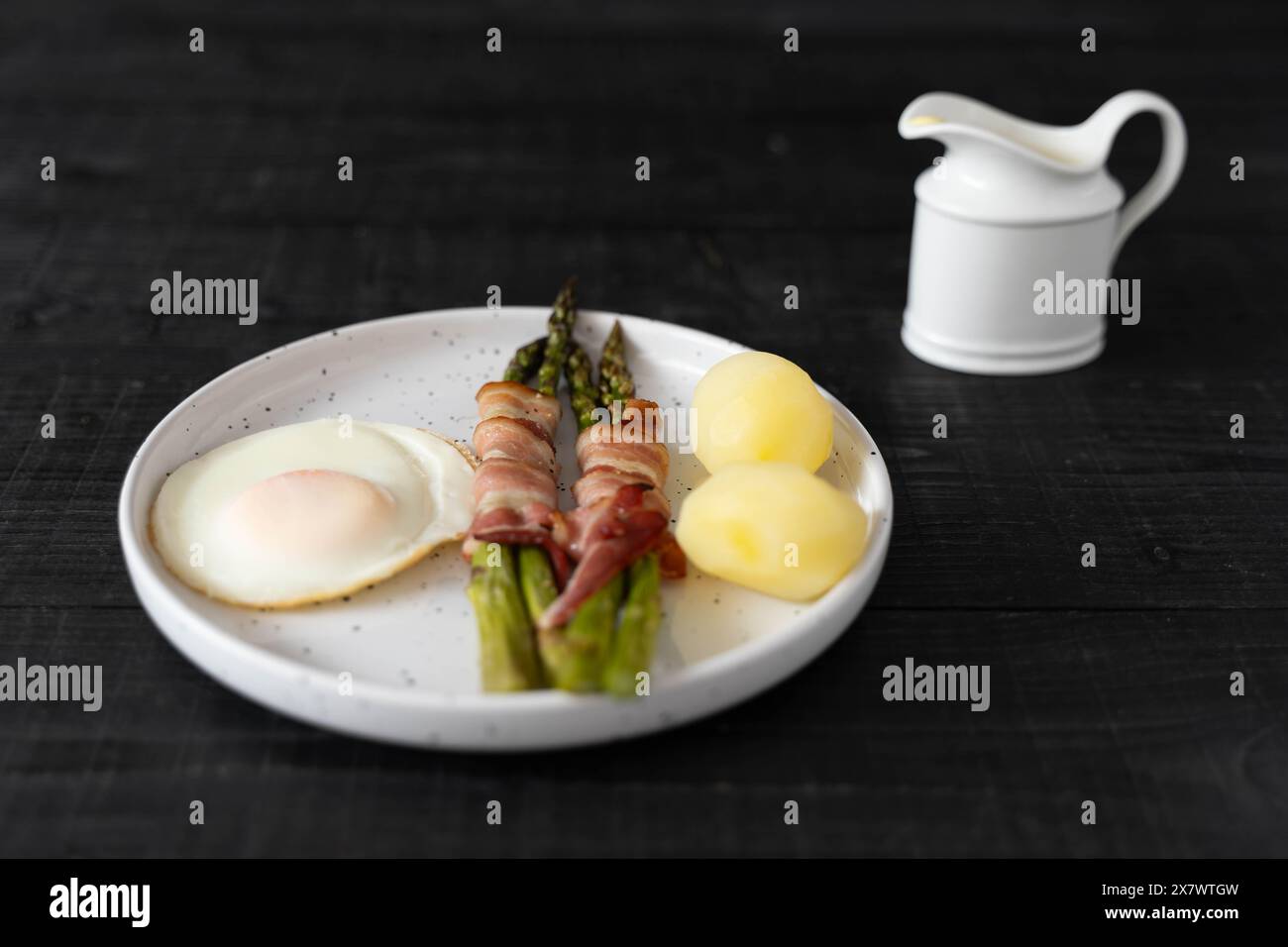 Asparagi verdi cotti al forno avvolti in pancetta e serviti con patate bollite e uova, su splendidi piatti bianchi su una superficie di legno nero Foto Stock