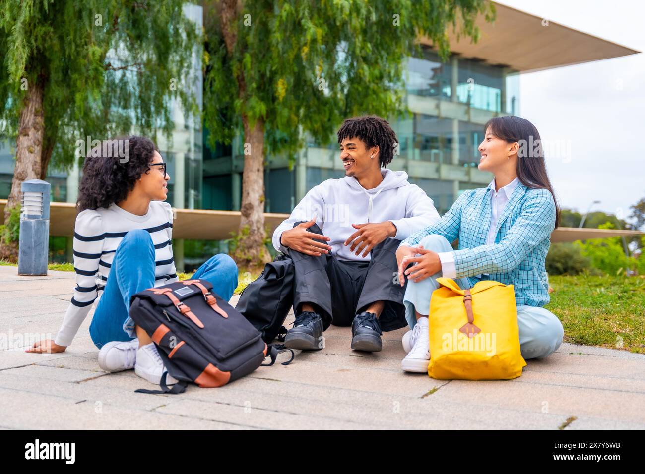 Foto a figura intera di tre studenti multirazziali sorridenti seduti e chiacchierati rilassati nel campus universitario Foto Stock