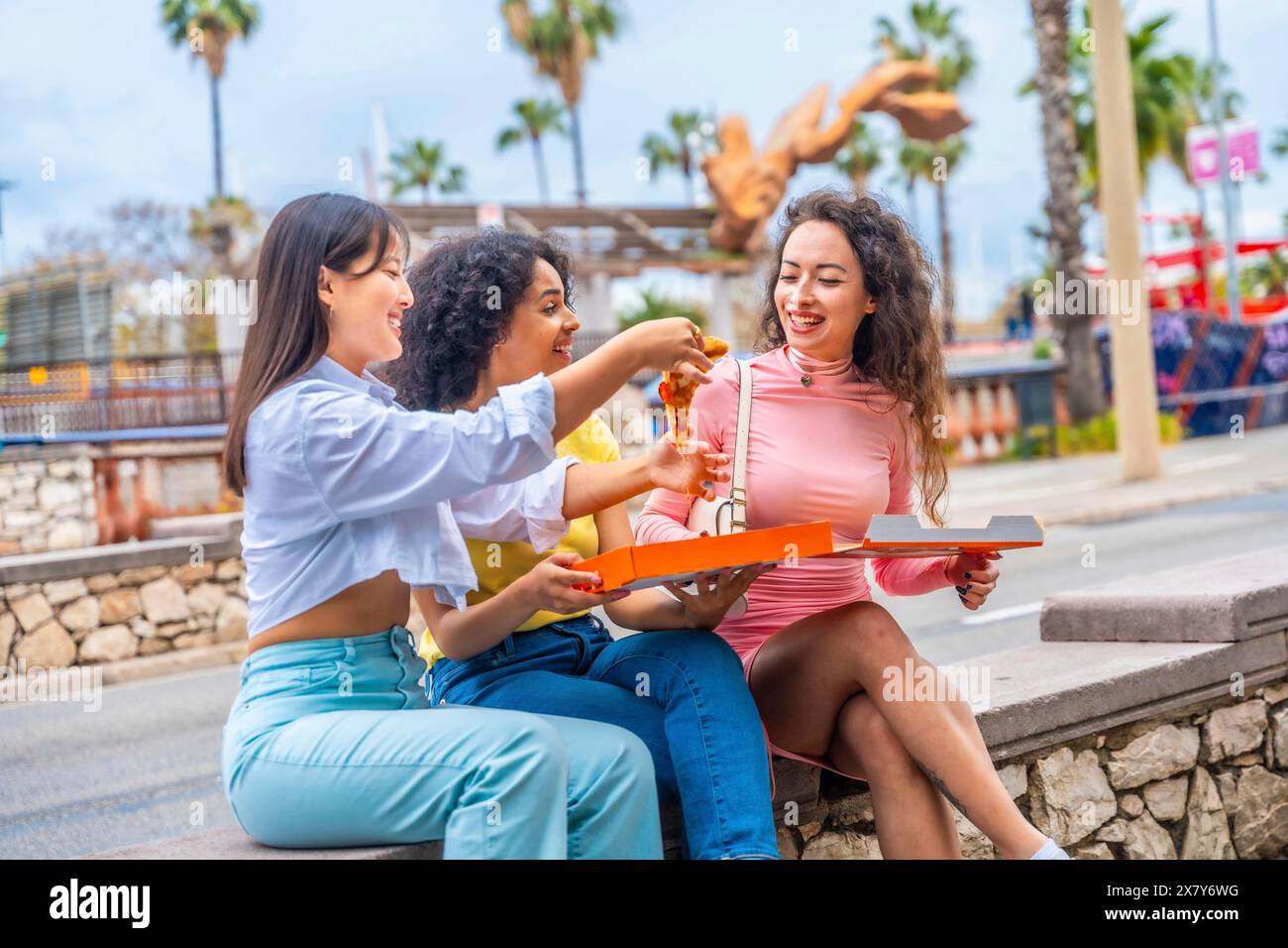 Tre amiche multietniche che mangiano pizza sedute in una panchina della città Foto Stock