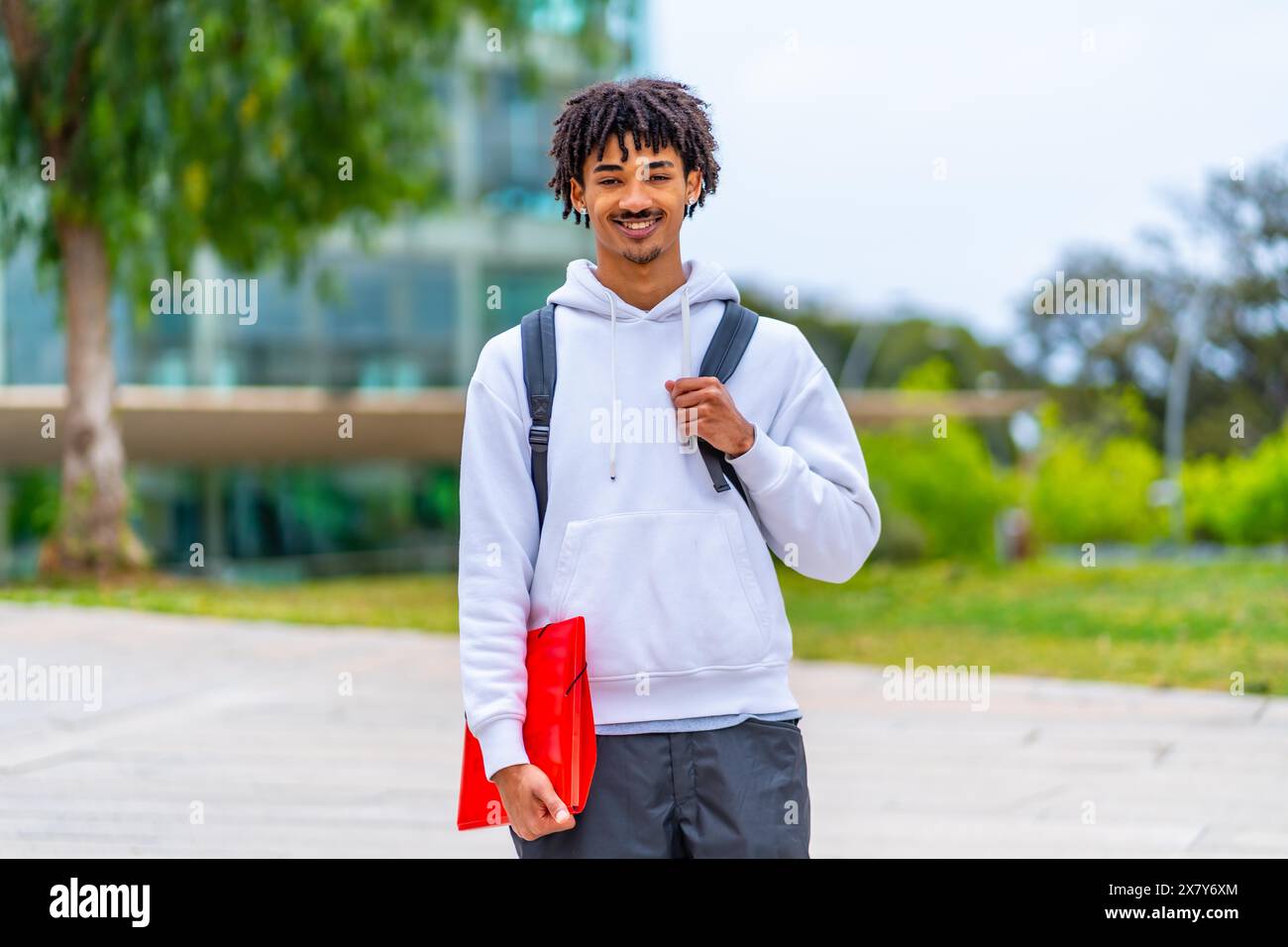 Ritratto di uno studente afro-americano in piedi nel campus sorridente alla macchina fotografica che tiene una cartella e porta uno zaino Foto Stock