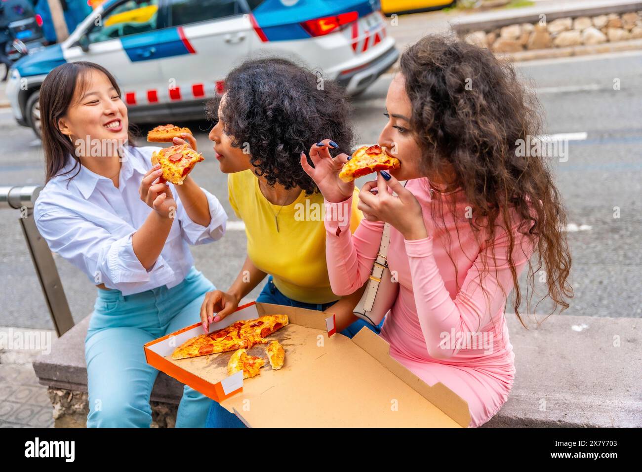 Tre donne multirazziali che condividono la pizza sedute insieme per strada Foto Stock