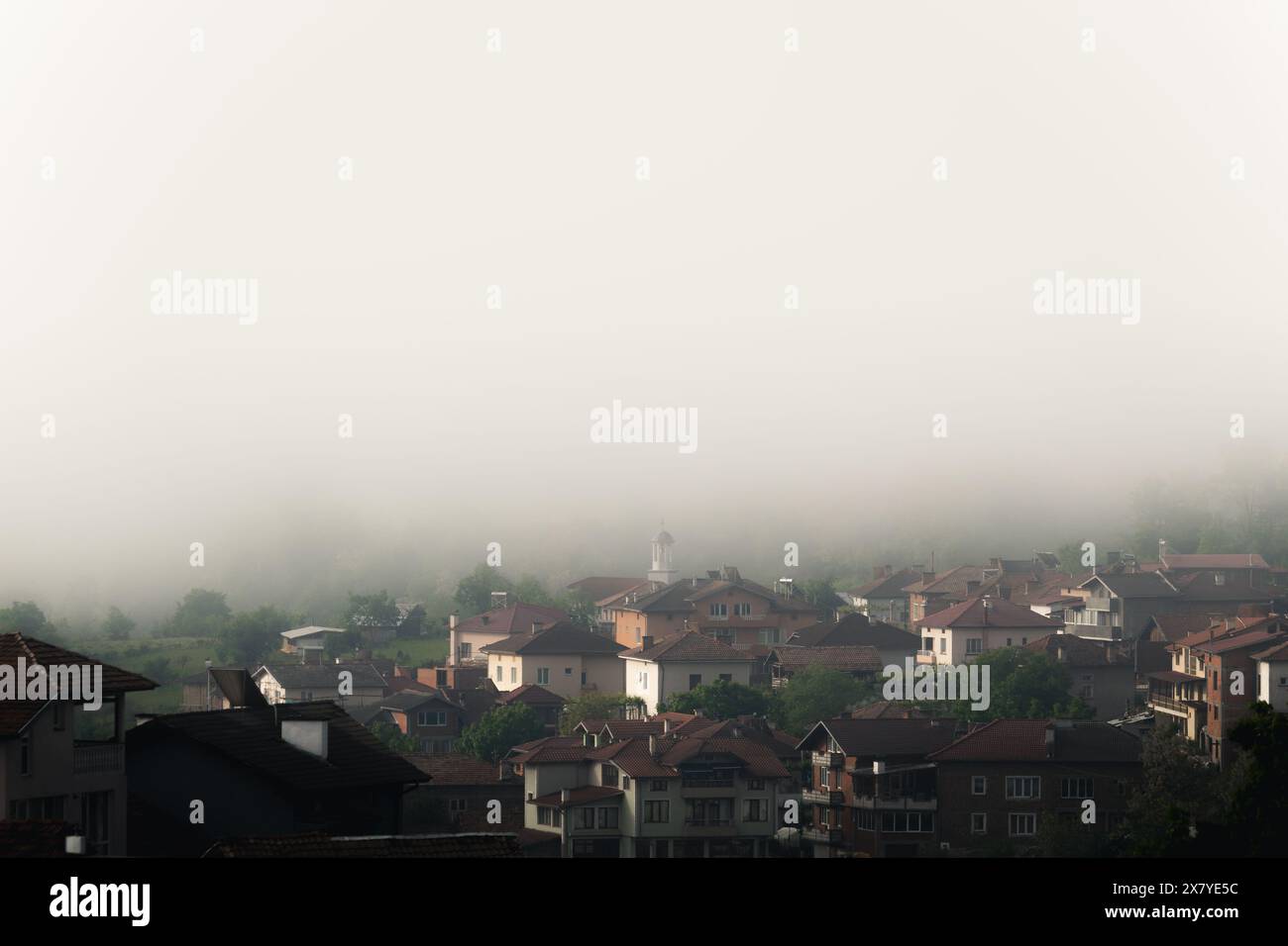 Bulgaria - 05 22 2024: Nebbia su un grande villaggio con una piccola chiesa bianca al centro sullo sfondo coperta di nebbia. Foto Stock