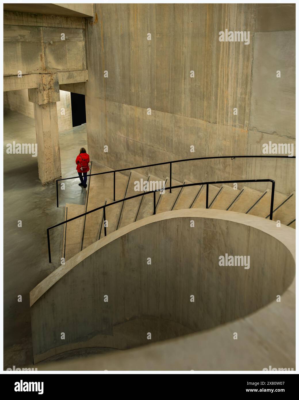 Scalinata modernistica a spirale in cemento con banister e una persona rivestita di rosso in fondo alla scala. Pareti in calcestruzzo lisce e rifinite Foto Stock