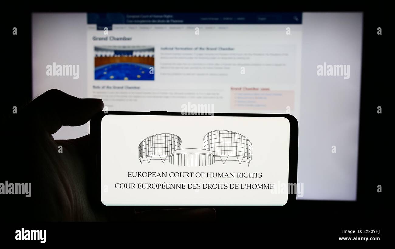 Persona che detiene un cellulare con il logo del tribunale internazionale della Corte europea dei diritti dell'uomo (CEDU) di fronte alla pagina web. Mettere a fuoco il display del telefono. Foto Stock