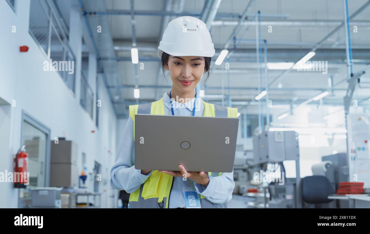 Una donna asiatica felice e sorridente ingegnere in bianco Hard Hat in piedi con computer portatile alla fabbrica di produzione elettronica. Dipendente di successo che si concentra sullo schermo del PC Foto Stock