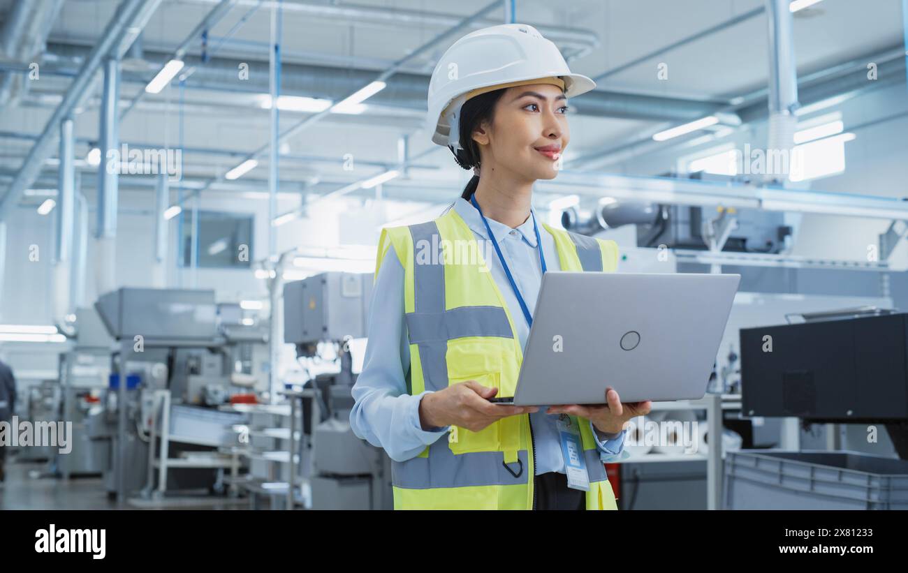 Una donna asiatica felice e sorridente ingegnere in bianco Hard Hat in piedi con computer portatile alla fabbrica di produzione elettronica. Dipendente di successo che si concentra sullo schermo del PC Foto Stock