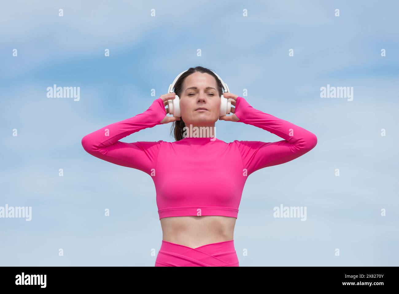 Donna sportiva che ascolta la musica, indossa abiti sportivi rosa contro il cielo blu Foto Stock