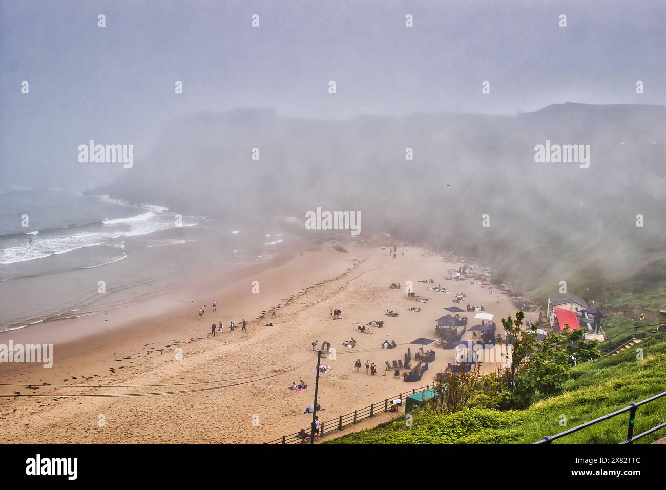 Una spiaggia nebbiosa con persone che si rilassano sulla sabbia e alcune tende allestite. Le onde oceaniche si schiantano dolcemente sulla riva, e lo sfondo lo è Foto Stock