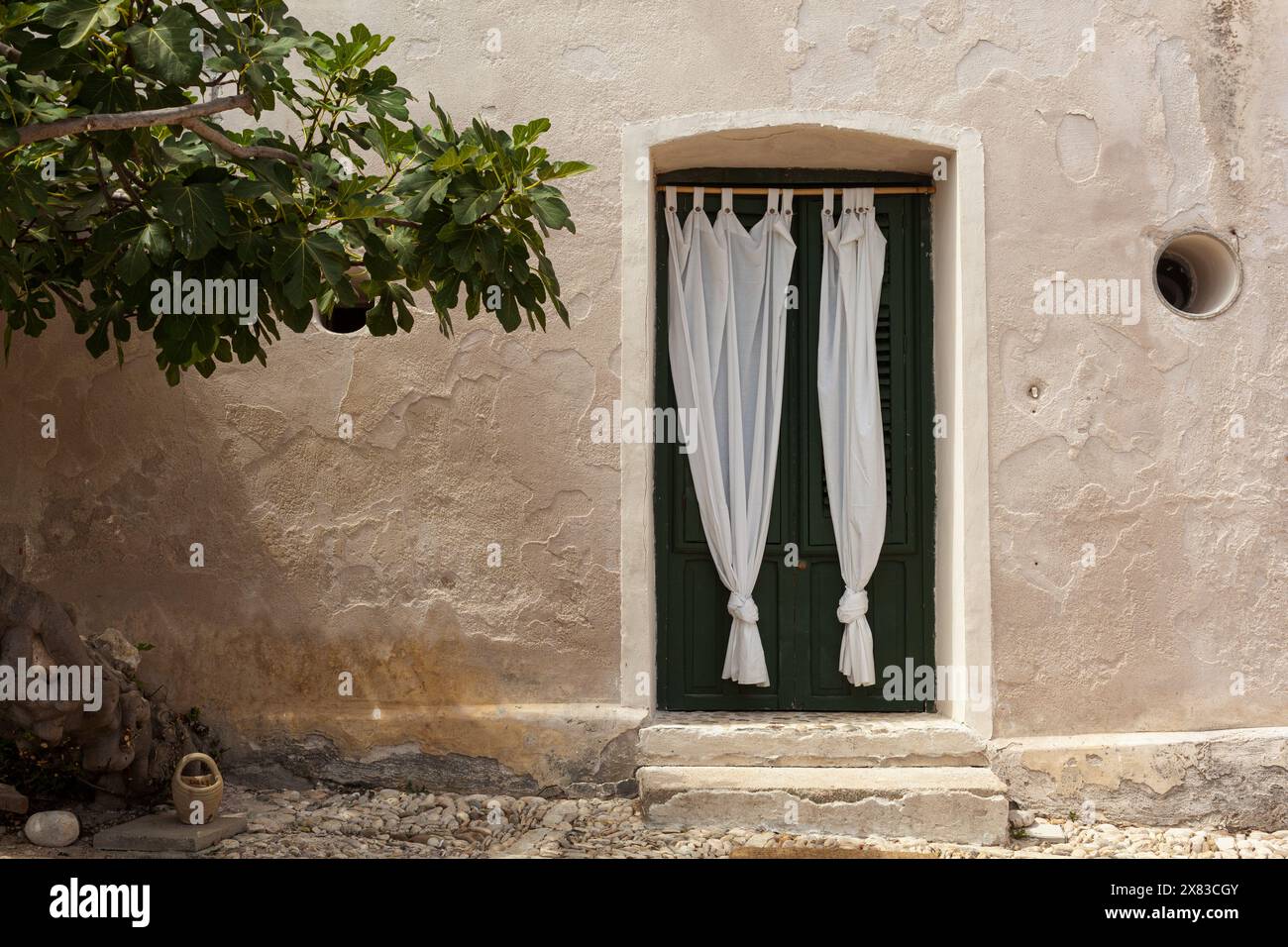 Vista di un'antica casa con tende bianche sulla porta, accanto a un fico secolare, Tonnara di Scopello, Sicilia. Foto Stock