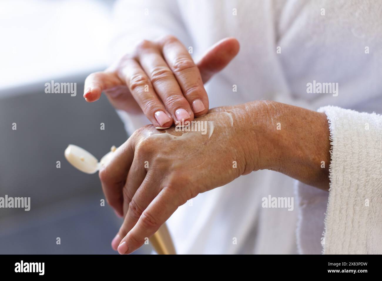 In bagno a casa, donna caucasica matura che indossa una vestaglia bianca, applicando lozione sulle mani Foto Stock