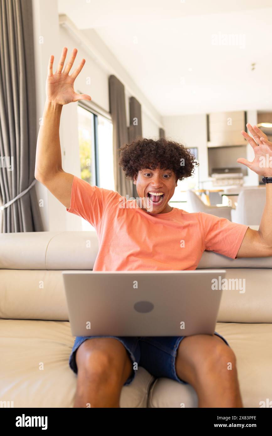 Un giovane maschio birazziale a casa, che usa il laptop sul divano, mostrando emozioni Foto Stock