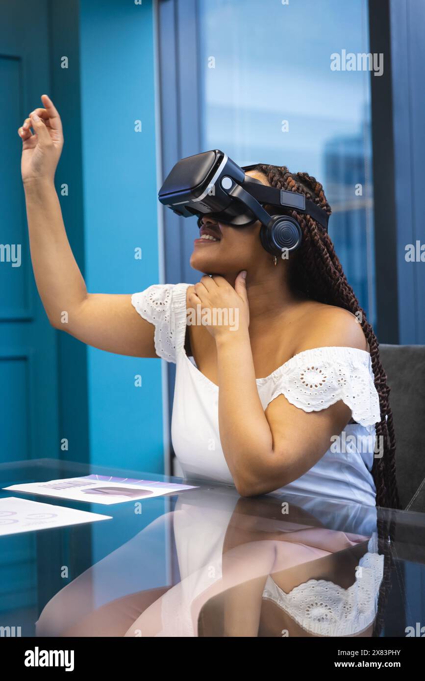 In un ufficio moderno, una giovane donna birazziale con trecce e spalle scoperte utilizza un visore VR Foto Stock