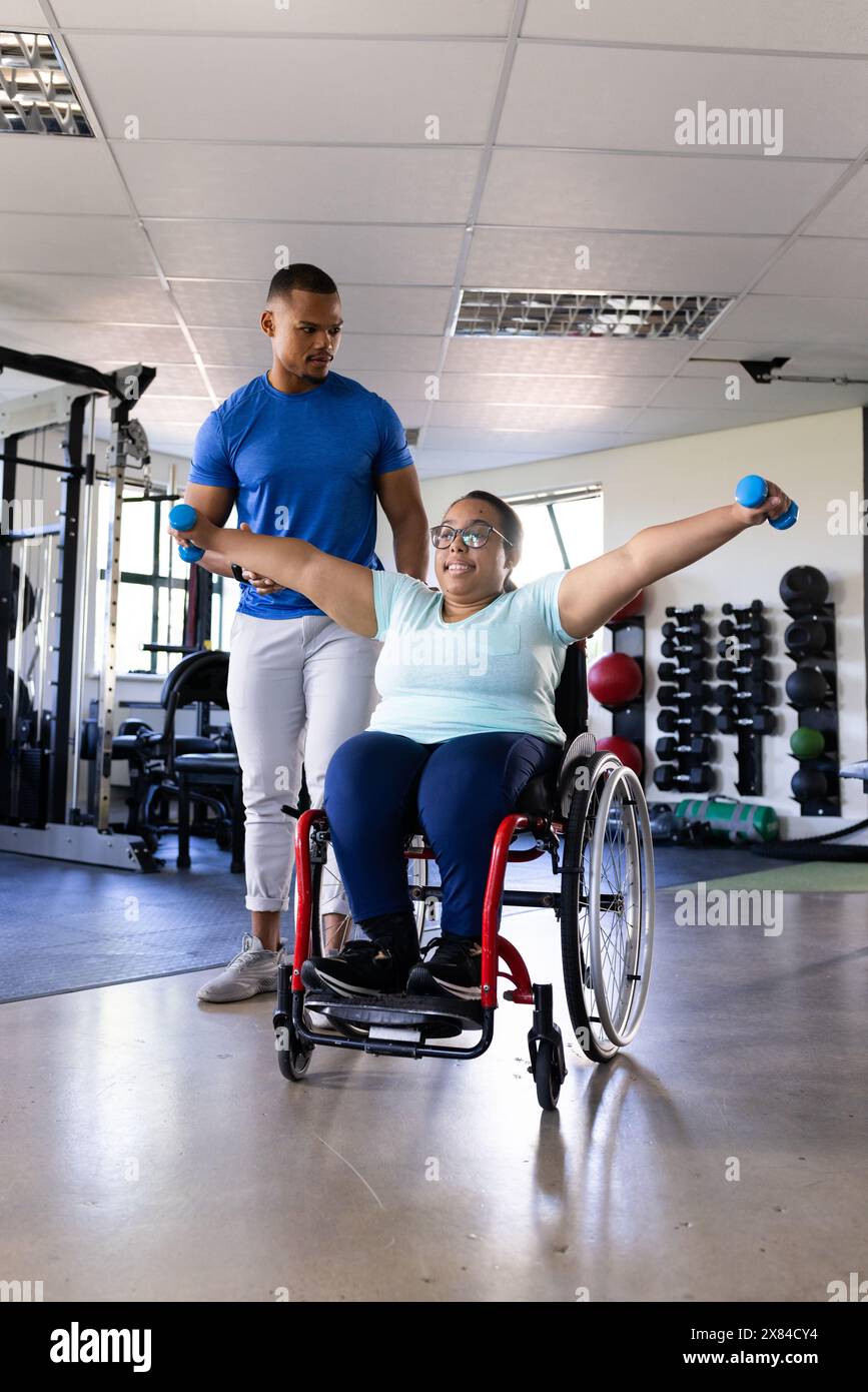 Al centro di riabilitazione, esercizi paraplegici femminili birazziali, mentre il terapista maschile solleva i pesi Foto Stock