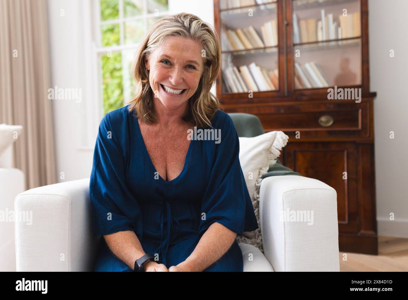 Donna anziana caucasica in abito blu che guarda le videochiamate a casa, sorridendo Foto Stock