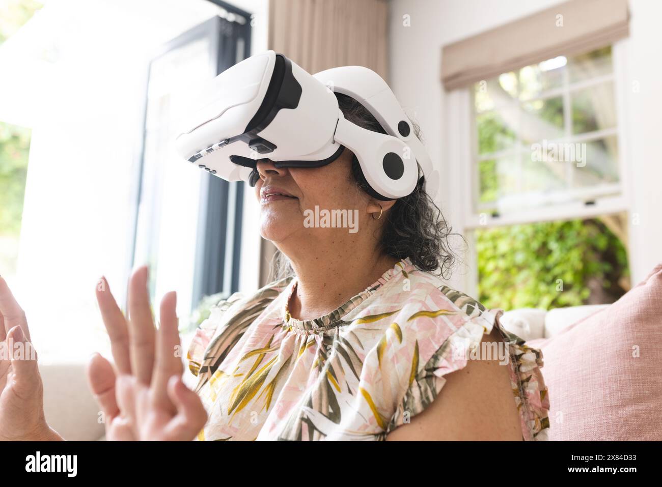 Una donna birazziale anziana che indossa un visore VR si siede sul divano di casa Foto Stock