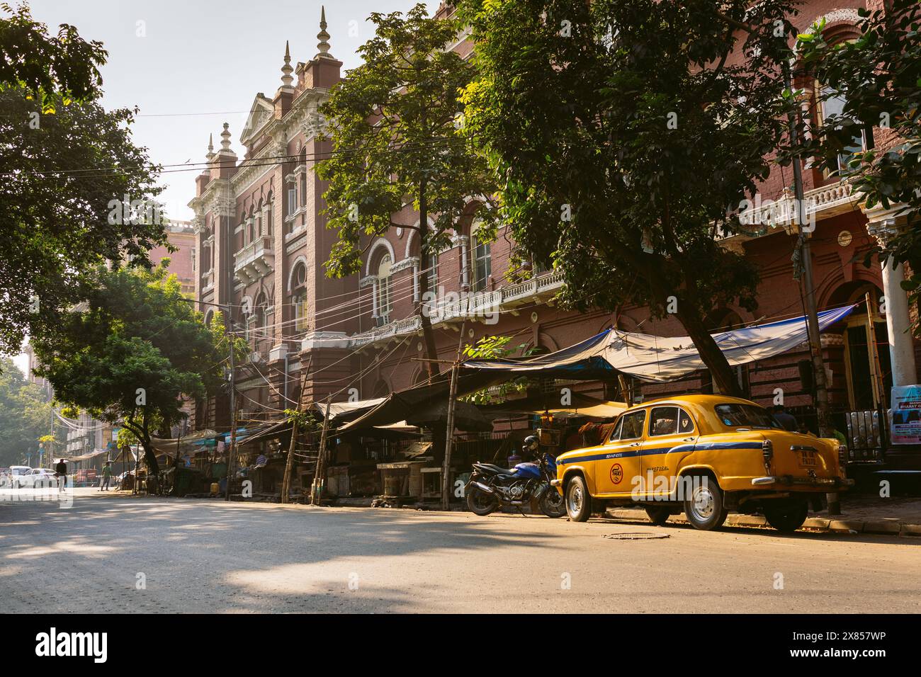 Kolkata, India - 20 ottobre 2023: Un tradizionale taxi indiano giallo è parcheggiato di fronte a edifici di architettura tradizionale con luce calda nel mor Foto Stock