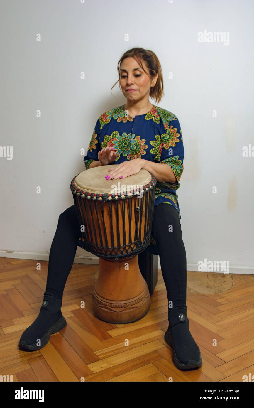 Una donna latina sola siede a casa con la sua batteria Yembe a fare musica. Sta praticando e studiando musica tradizionale africana e indossa un colorato Foto Stock