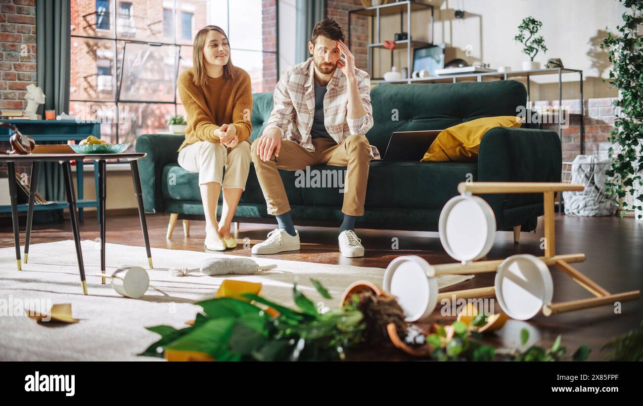 Momento divertente: Fiore in vaso rovinato da Dog dopo averlo rovesciato e aver fatto disordine in tutto l'appartamento. Coppia seduta sul divano con un aspetto di incredulità, frustrazione. Donna che sorride Foto Stock