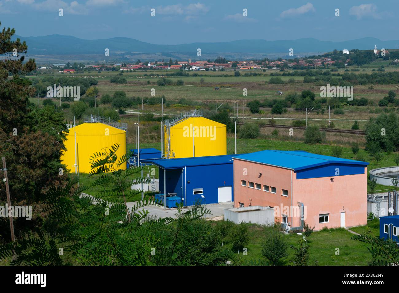 Una struttura industriale rurale che presenta cisterne gialle luminose, strutture blu e colline verdi ondulate con un villaggio lontano. Foto Stock