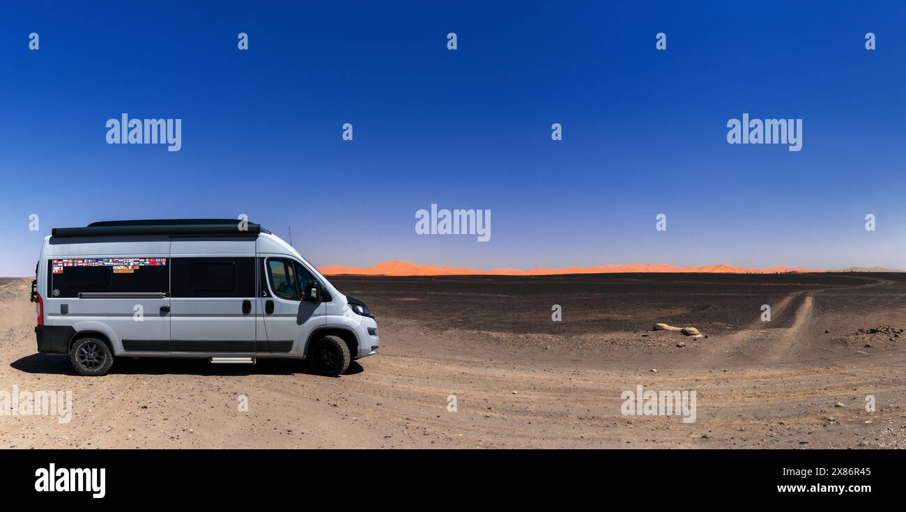 Merzouga, Marocco - 8 marzo 2024: Camper ben viaggiato parcheggiato in un paesaggio desolato con dune di sabbia arancione e deserto di roccia nera nel sud-est Foto Stock