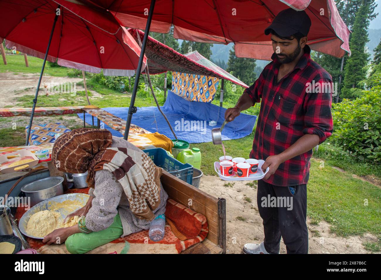 Una donna del Kashmir prepara tortillas di farina di mais mentre suo marito serve il Noon Chai (tè salato) ai turisti e alla gente del posto passando per la loro bancarella lungo la strada Doodhpathri nel distretto di Budgam, a sud-ovest di Srinagar. Le autorità hanno chiesto ai proprietari dei venditori di tè di liberare lo spazio che hanno occupato "illegalmente" entro dieci giorni o di affrontare azioni. I funzionari hanno detto a un'agenzia di stampa locale che l'autorità per lo sviluppo del turismo ha ricevuto molte lamentele per il fatto che i proprietari delle bancarelle di tè hanno tagliato rami di alberi, danneggiando così i pittoreschi prati. Sulla strada per i prati di Foto Stock