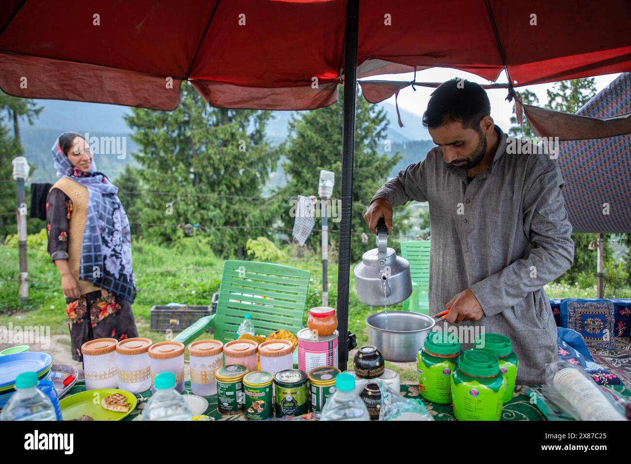 Un uomo del Kashmir prepara il Noon Chai (tè salato) mentre sua moglie aspetta di servirlo con tortillas di farina di mais ai turisti e alla gente del posto passando per la loro bancarella lungo la strada Doodhpathri nel distretto di Budgam, a sud-ovest di Srinagar. Le autorità hanno chiesto ai proprietari dei venditori di tè di liberare lo spazio che hanno occupato "illegalmente" entro dieci giorni o di affrontare azioni. I funzionari hanno detto a un'agenzia di stampa locale che l'autorità per lo sviluppo del turismo ha ricevuto molte lamentele per il fatto che i proprietari delle bancarelle di tè hanno tagliato rami di alberi, danneggiando così i pittoreschi prati. Sulla strada per me Foto Stock