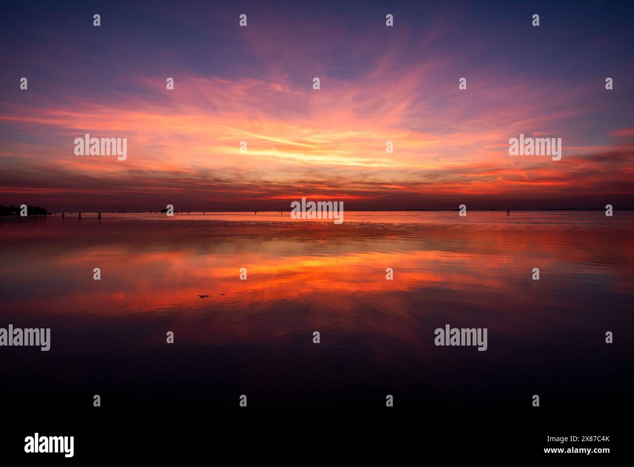 Il tramonto fiammeggiante con colori rosso e arancione si riflette sulle calme acque della laguna veneziana con mattoni e alberi fotografati dal Lido di VE Foto Stock