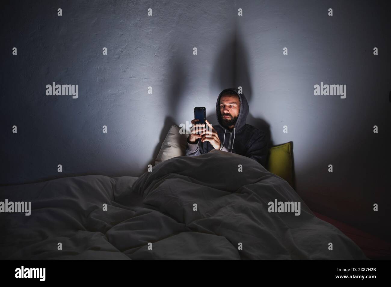 Uomo che utilizza uno smartphone seduto sul letto in una stanza buia Foto Stock