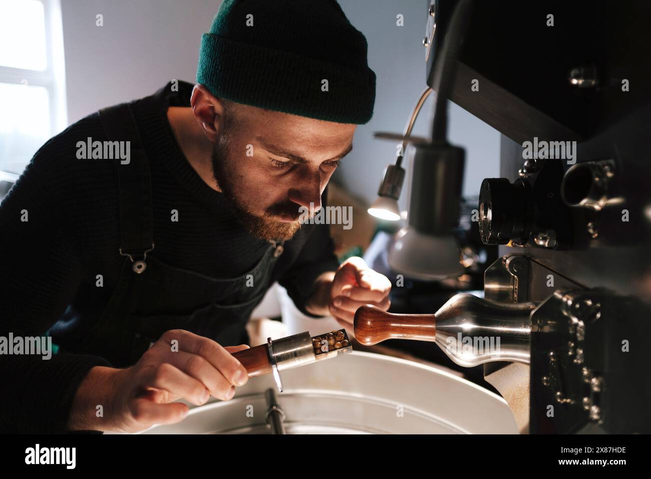 Tostapane con cappello in maglia e esame dei chicchi di caffè vicino a macchinari Foto Stock