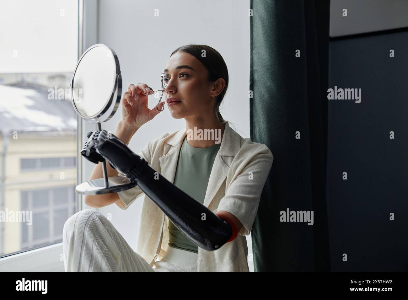 Giovane donna con braccio bionico che guarda se stessa allo specchio usando un arricciacapelli ciglia Foto Stock