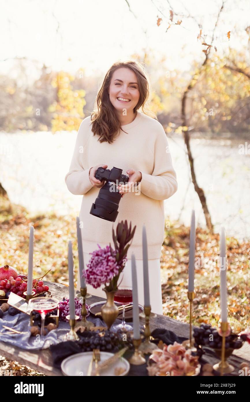 Donna sorridente che tiene la macchina fotografica vicino a un tavolo decorato nella foresta Foto Stock