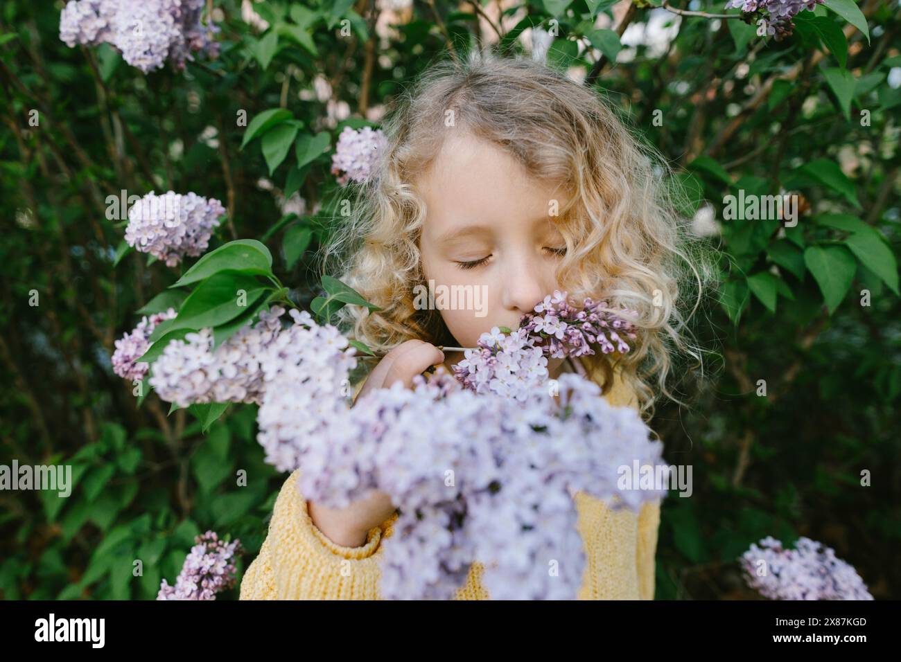Bionda ragazza dai capelli ricci che odora lills vicino alle piante Foto Stock