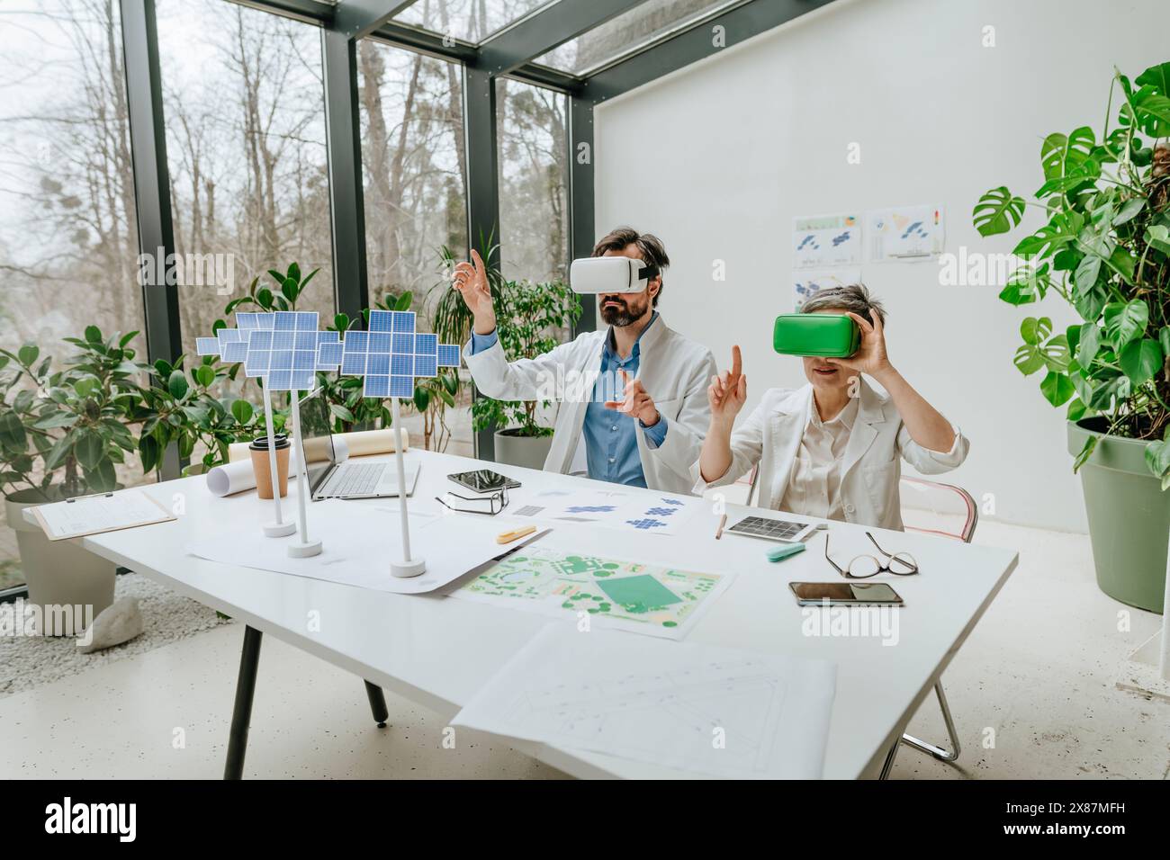 Ingegneri che indossano simulatori di realtà virtuale e lavorano a progetti di pannelli solari alla scrivania Foto Stock