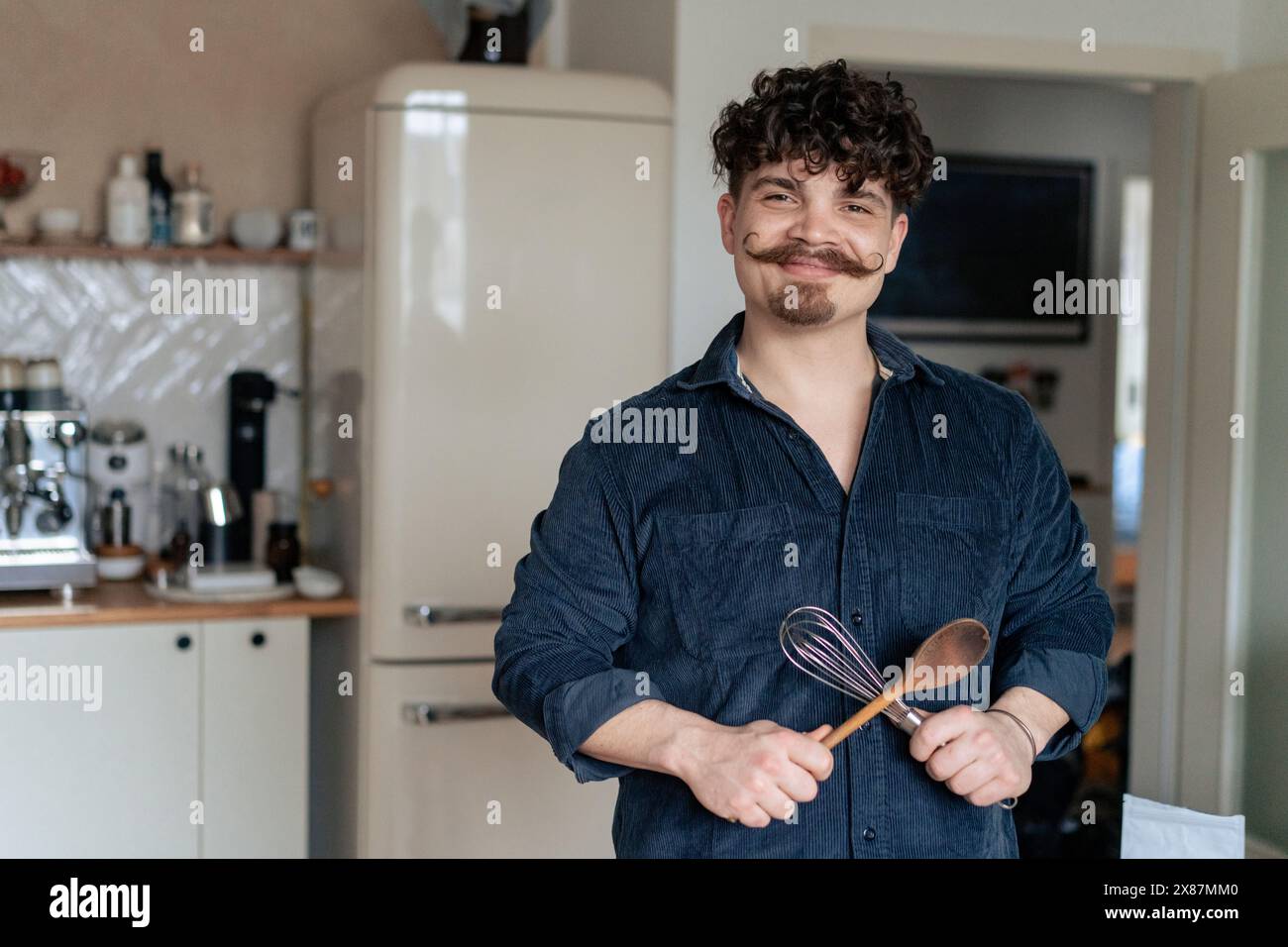 Uomo sorridente che tiene in mano un cucchiaio di legno e una frusta in cucina a casa Foto Stock