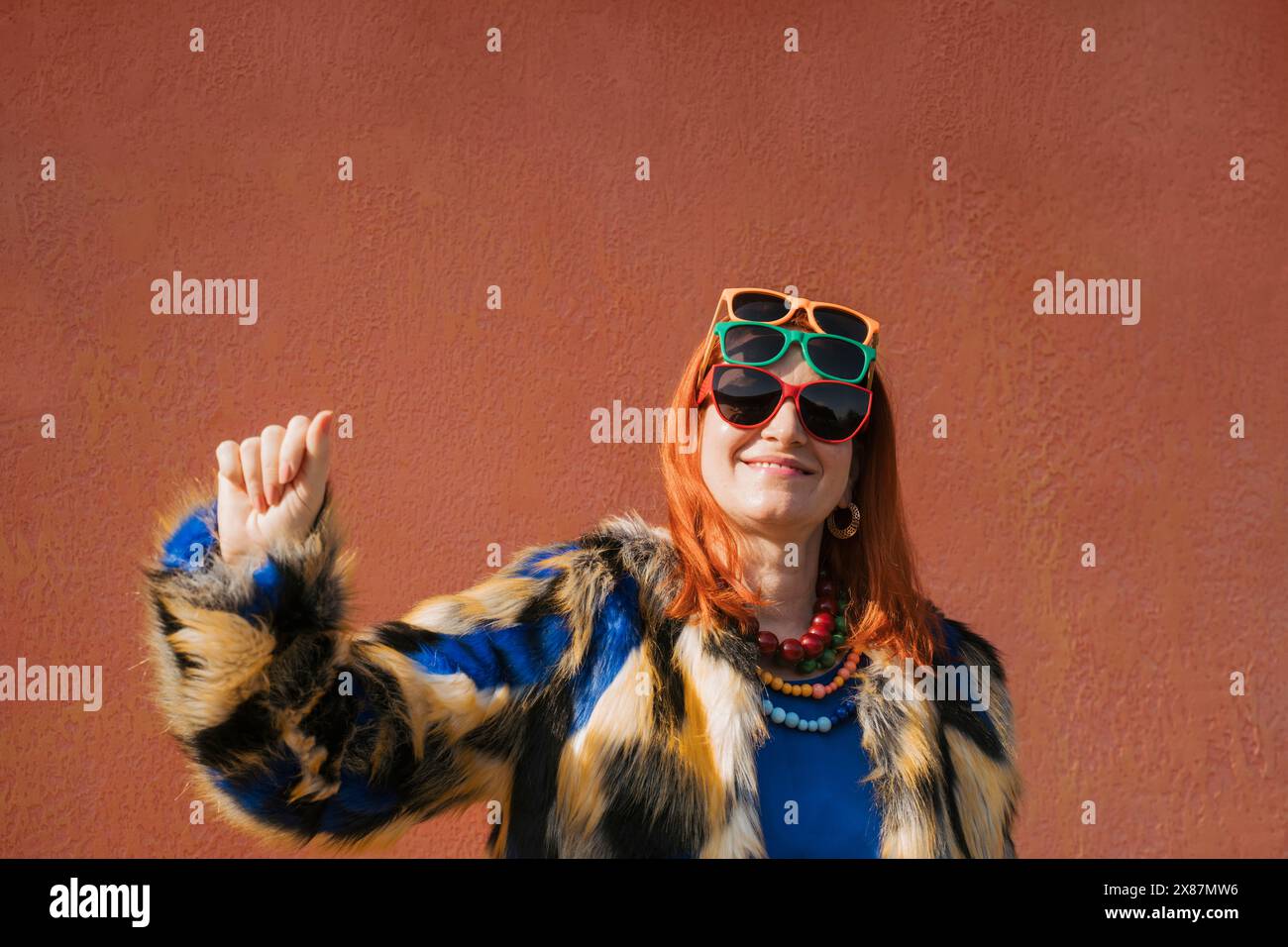 Donna alla moda che indossa occhiali da sole di tre colori in piedi davanti al muro marrone Foto Stock
