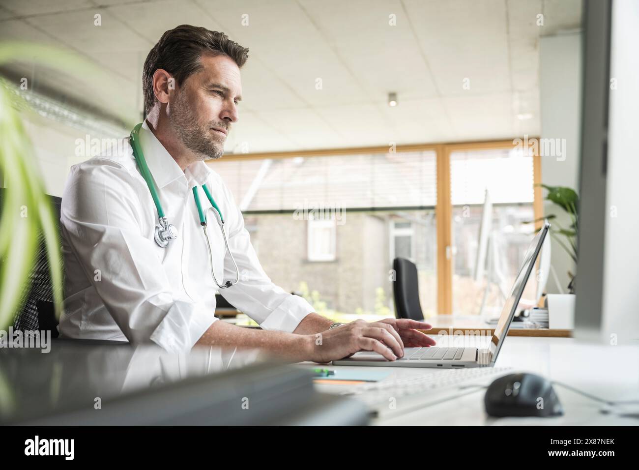 Medico maturo che usa un computer portatile seduto alla scrivania in ufficio Foto Stock