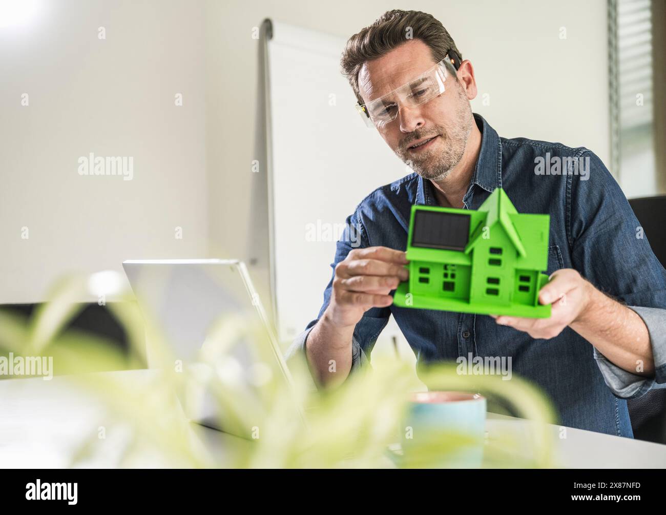 Architetto maturo che indossa occhiali intelligenti che esamina la casa modello alla scrivania in ufficio Foto Stock