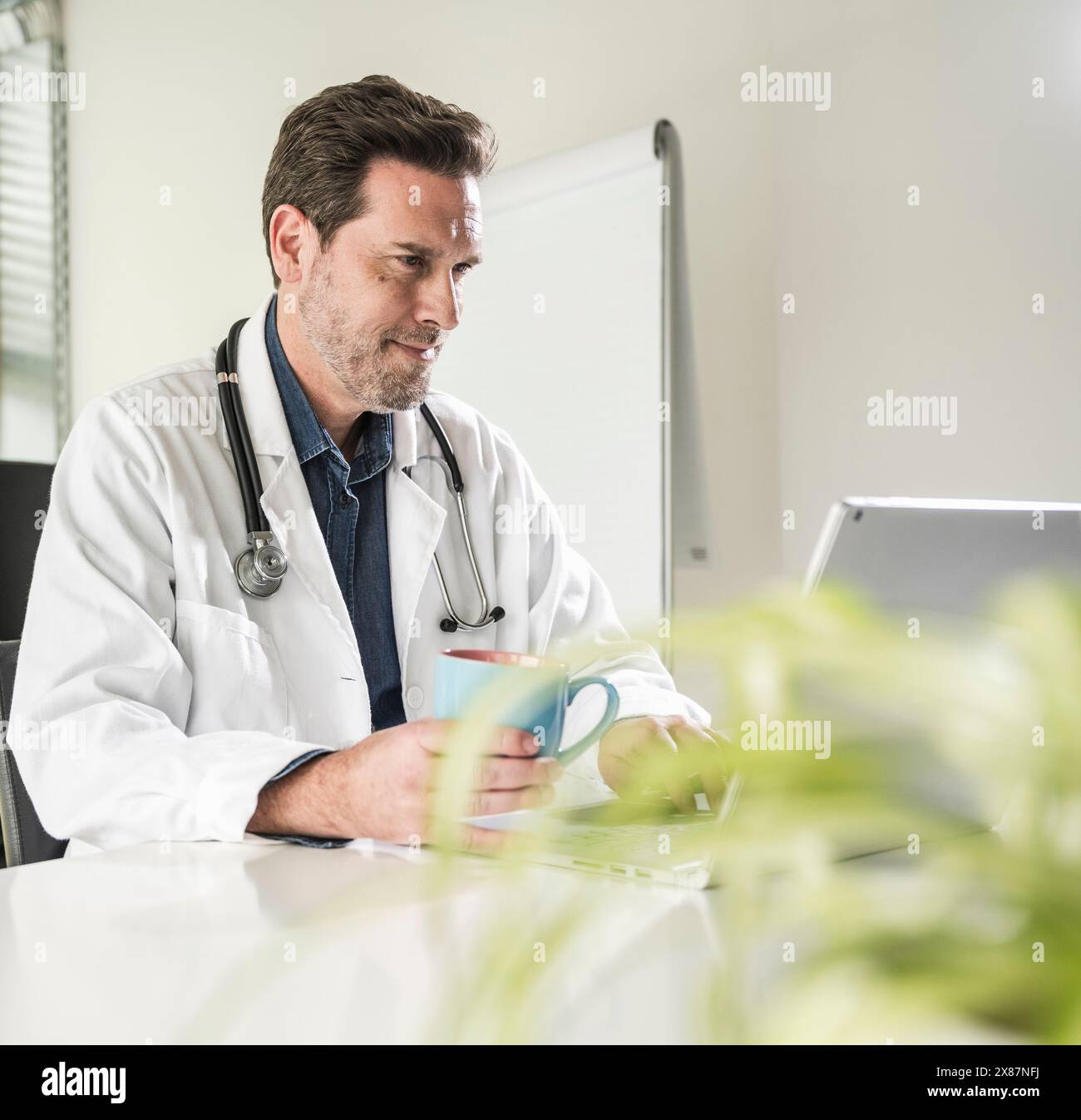 Medico sicuro seduto con una tazza di caffè utilizzando un computer portatile alla scrivania dell'ufficio Foto Stock