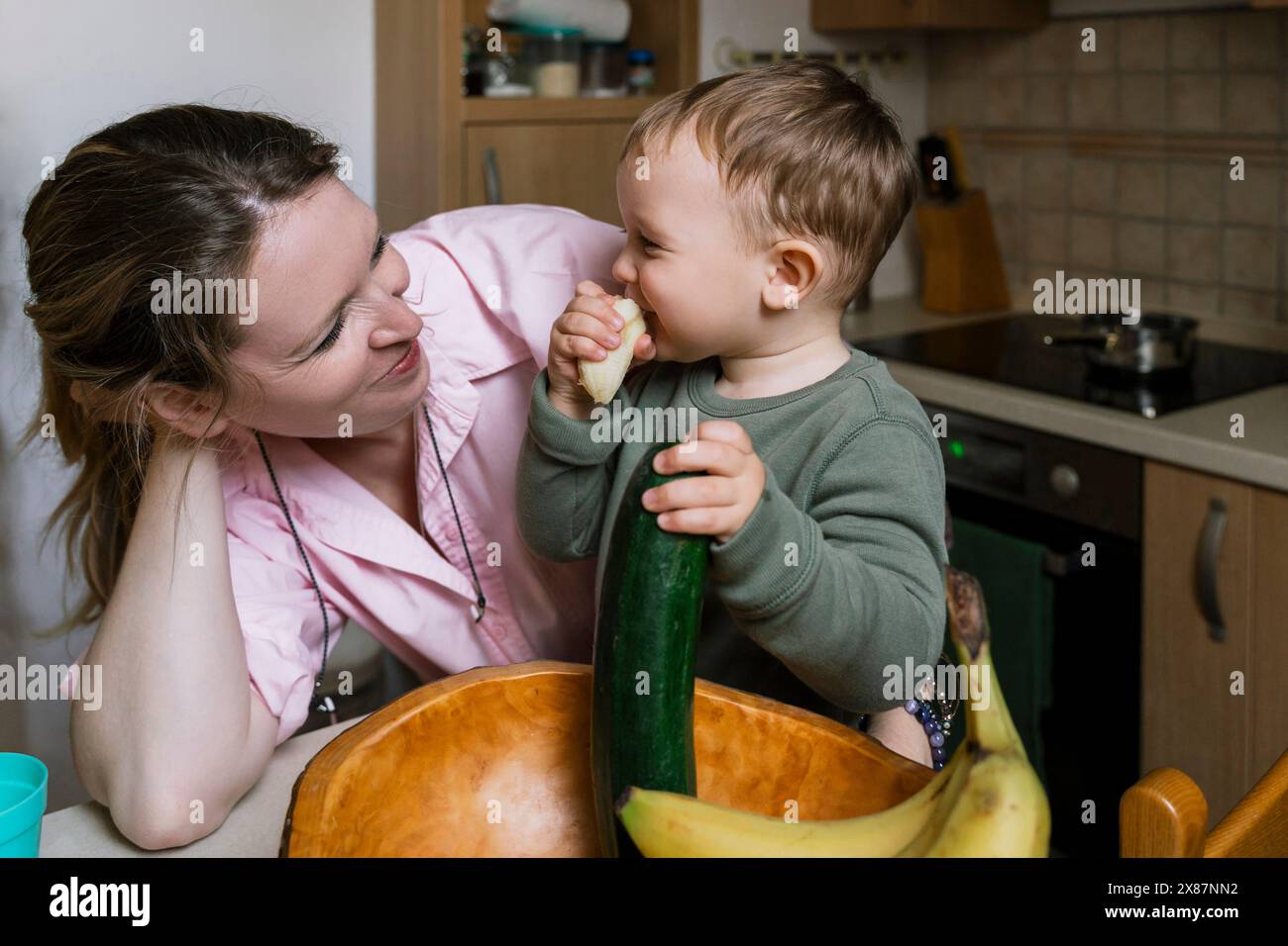 Madre sorridente che guarda un ragazzo che mangia cetriolo in cucina Foto Stock