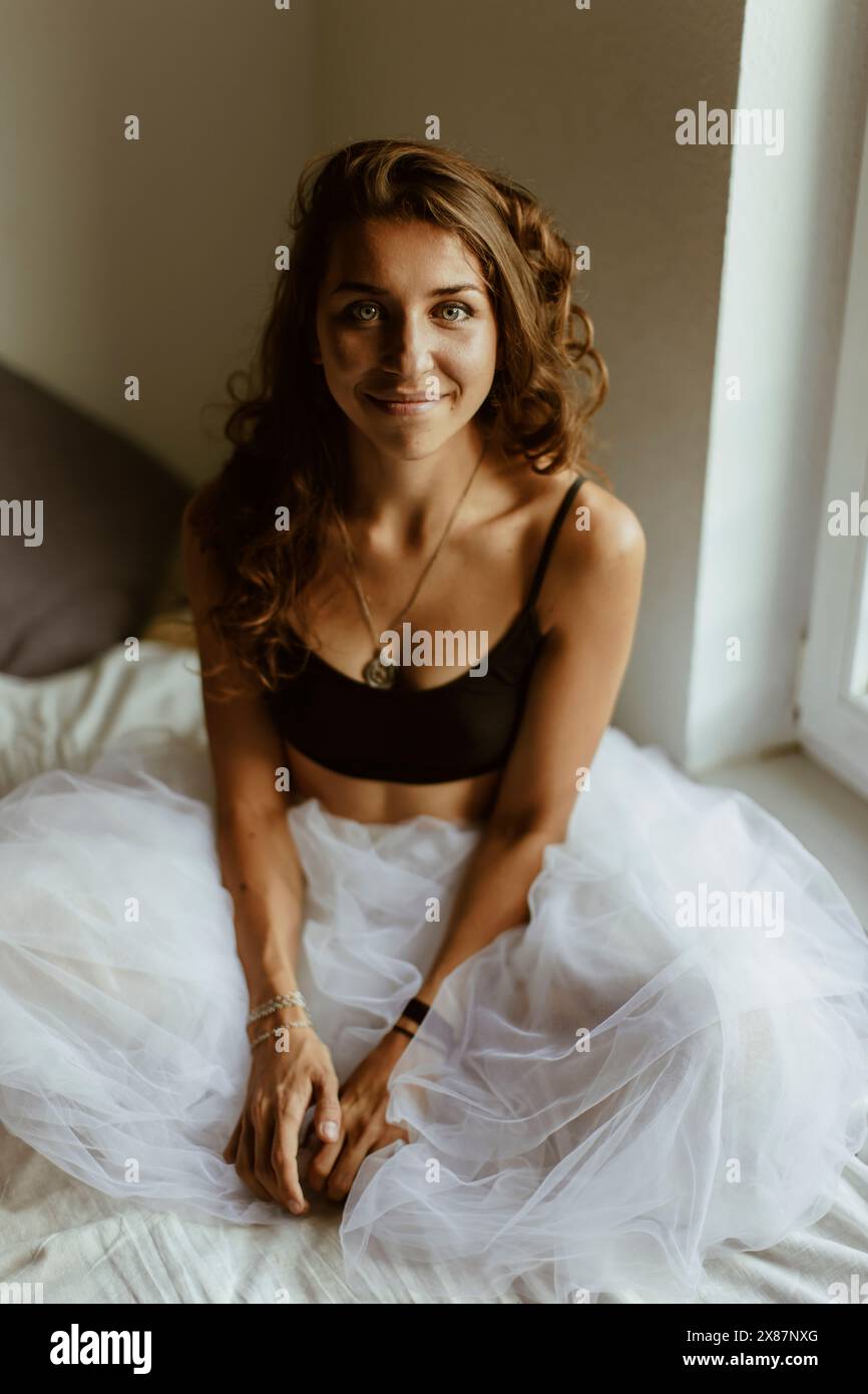 Giovane donna sorridente con capelli marroni seduta sul letto a casa Foto Stock