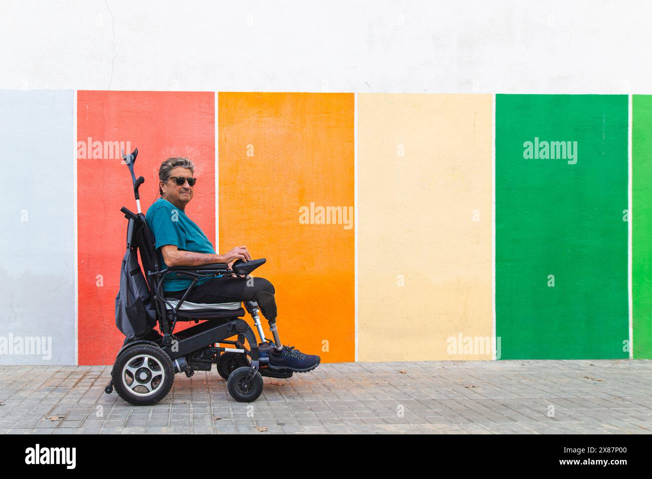 Uomo anziano con disabilità in sedia a rotelle motorizzata accanto a una parete colorata Foto Stock
