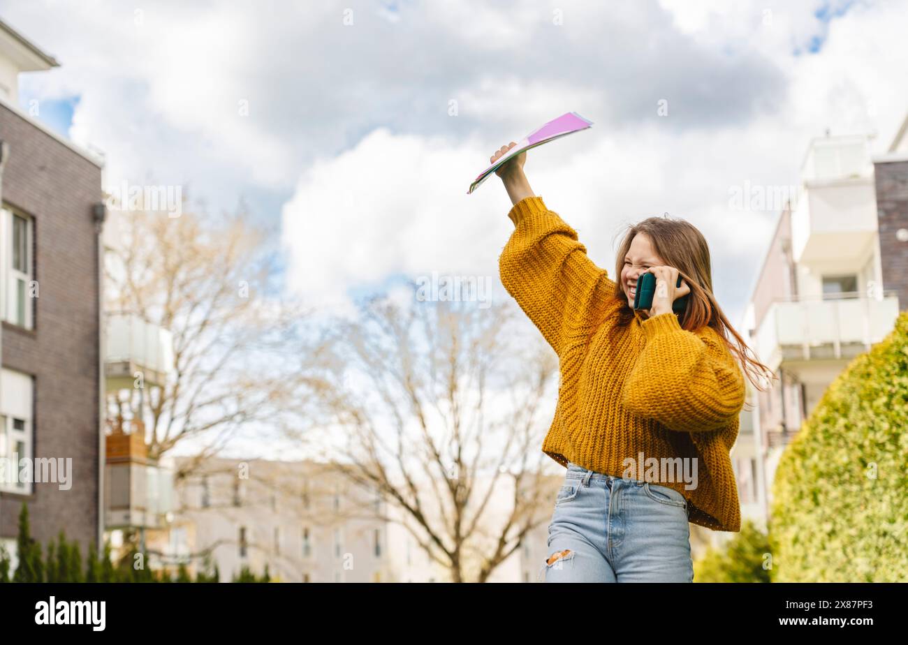 Allegra ragazza che parla con uno smartphone e tiene in mano le cartelle sotto il cielo nuvoloso Foto Stock