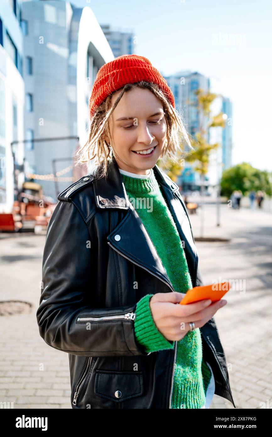 Giovane donna sorridente che indossa una giacca in pelle utilizzando il telefono cellulare nelle giornate di sole Foto Stock