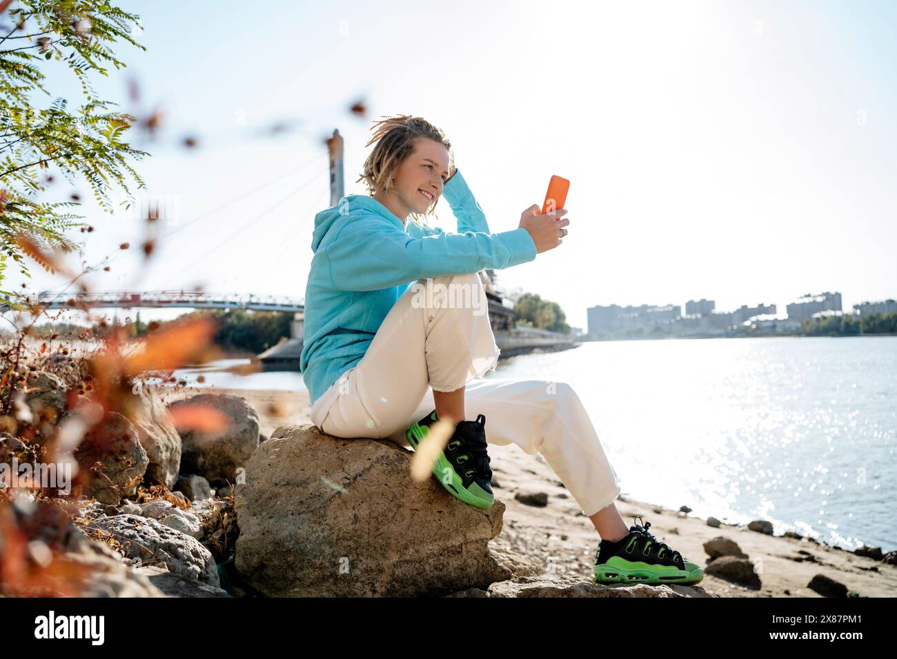Donna sorridente che scatta selfie con uno smartphone seduto sulla roccia sulla riva del fiume Foto Stock