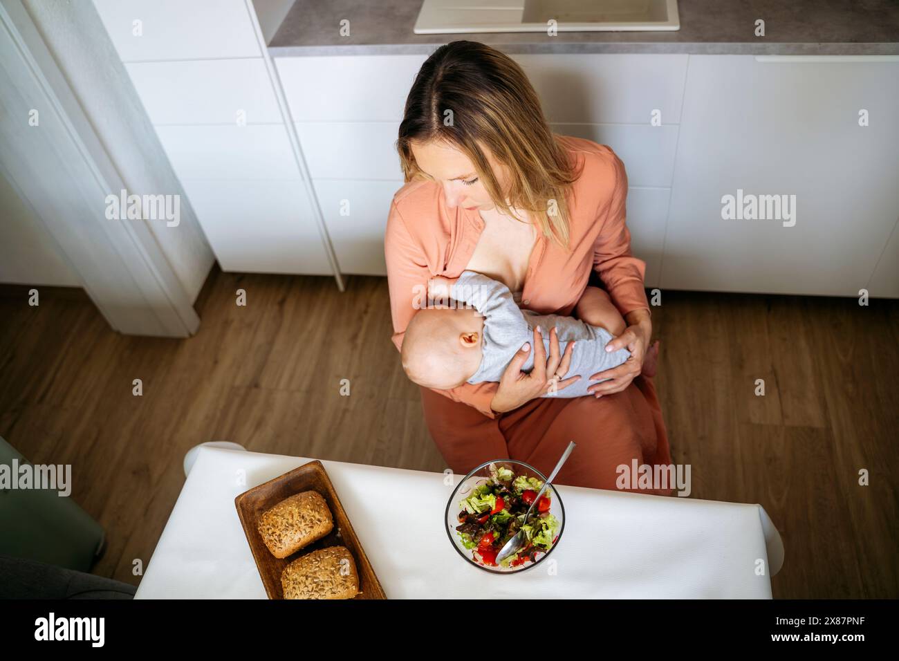 Donna bionda che allatta il bambino seduto con l'insalata al tavolo da pranzo in cucina Foto Stock