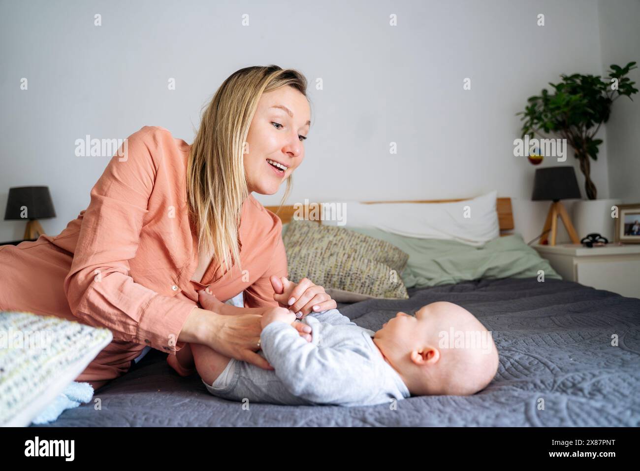 Madre bionda sorridente che gioca con il figlio del bambino sdraiato sul letto di casa Foto Stock