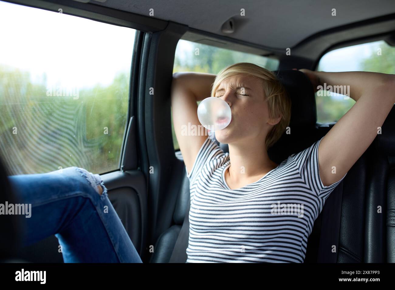 Giovane donna bionda con gli occhi chiusi che soffiano gomma da bolla seduta in auto Foto Stock
