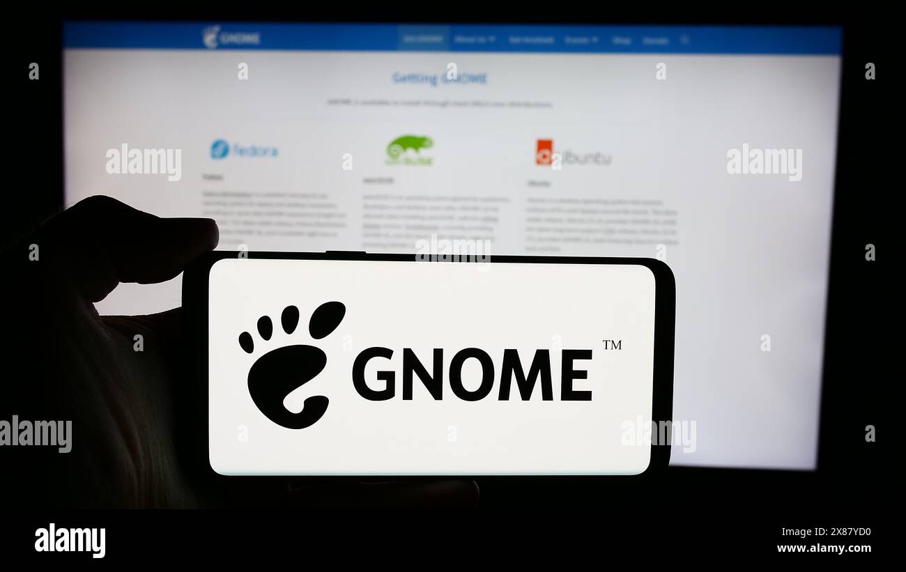 Persona che detiene un cellulare con il logo open source GNU Network Object Model Environment (GNOME) davanti alla pagina web. Mettere a fuoco il display del telefono. Foto Stock