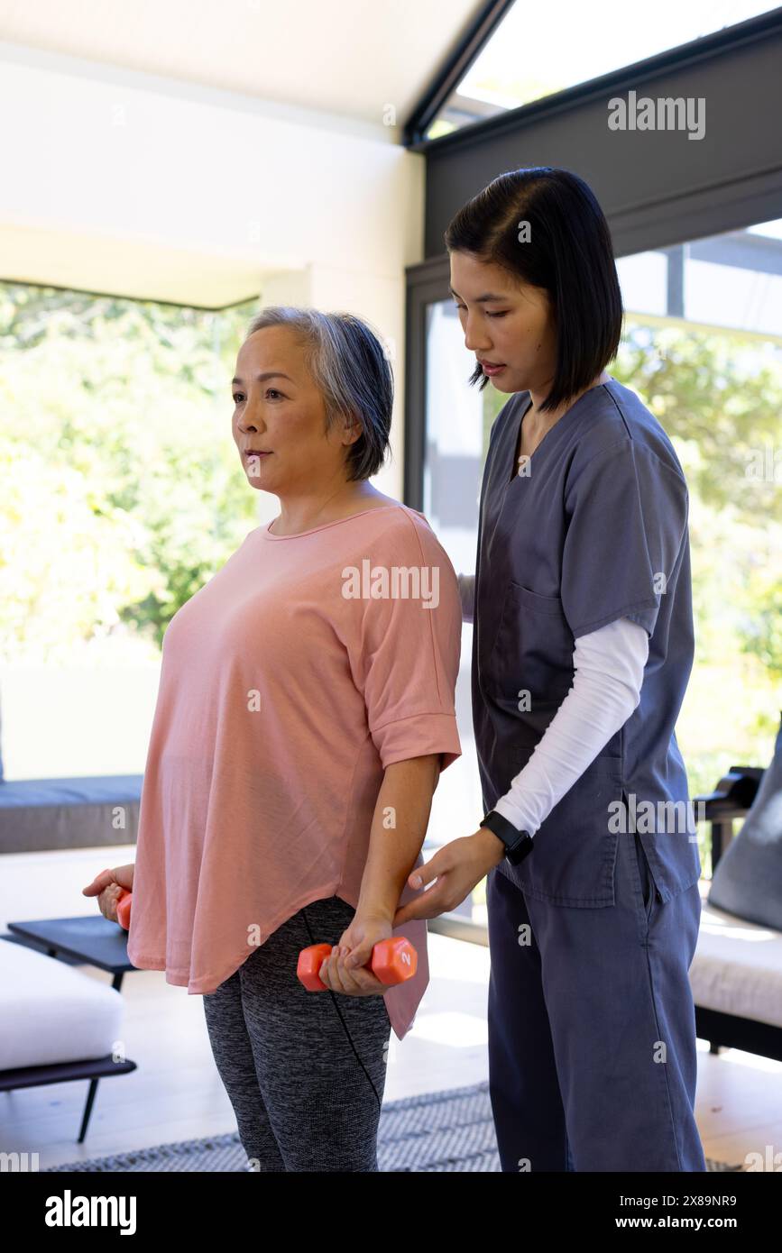 A casa, giovane infermiera asiatica e paziente di mezza età che solleva pesi Foto Stock