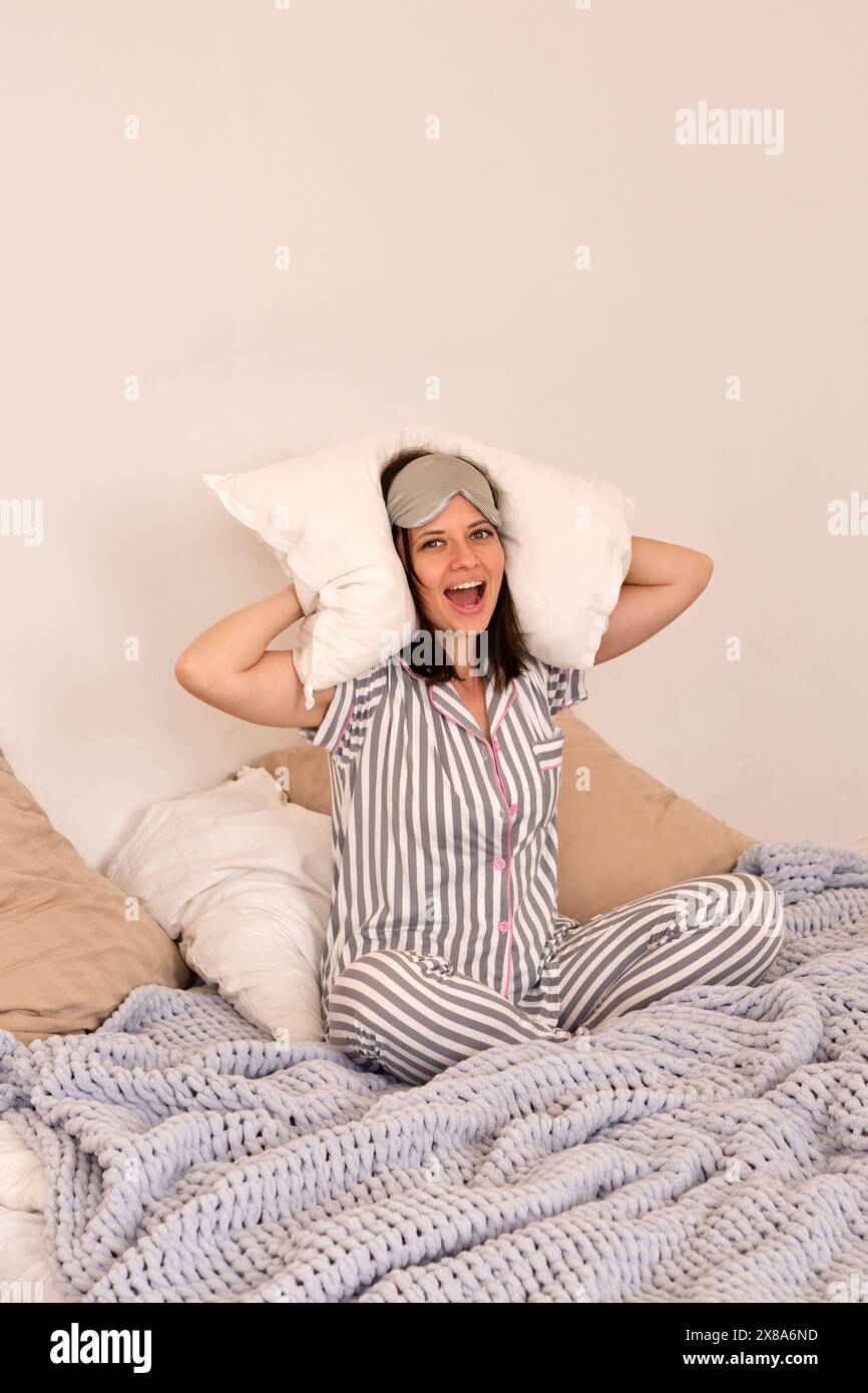 Donna in pigiama a righe grigie e una maschera dormiente che ride con cuscini dietro la testa. Foto Stock